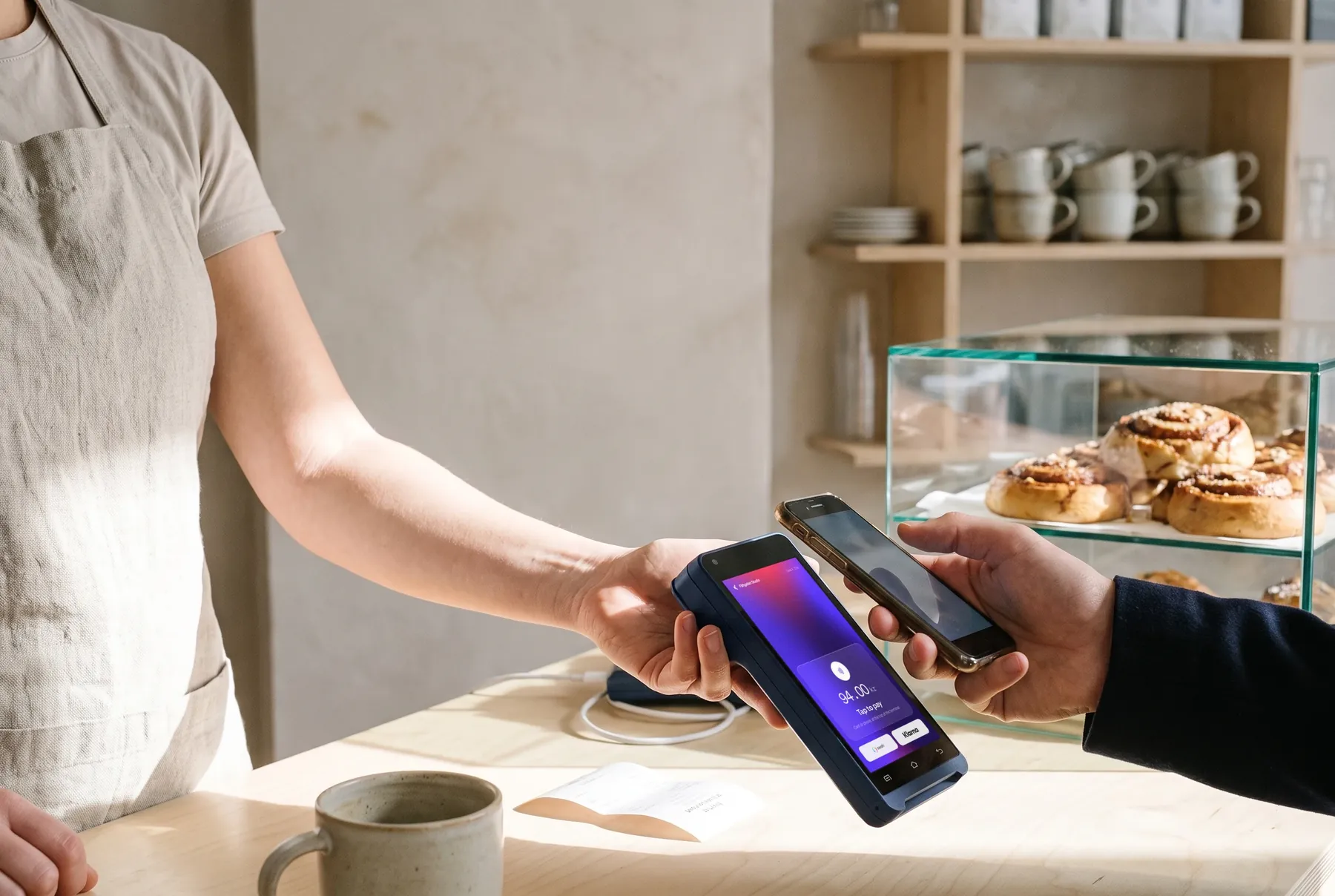 A barista holds a Karo Pay terminal across a café counter and a customer taps it with their phone, cinnamon buns in the case behind.